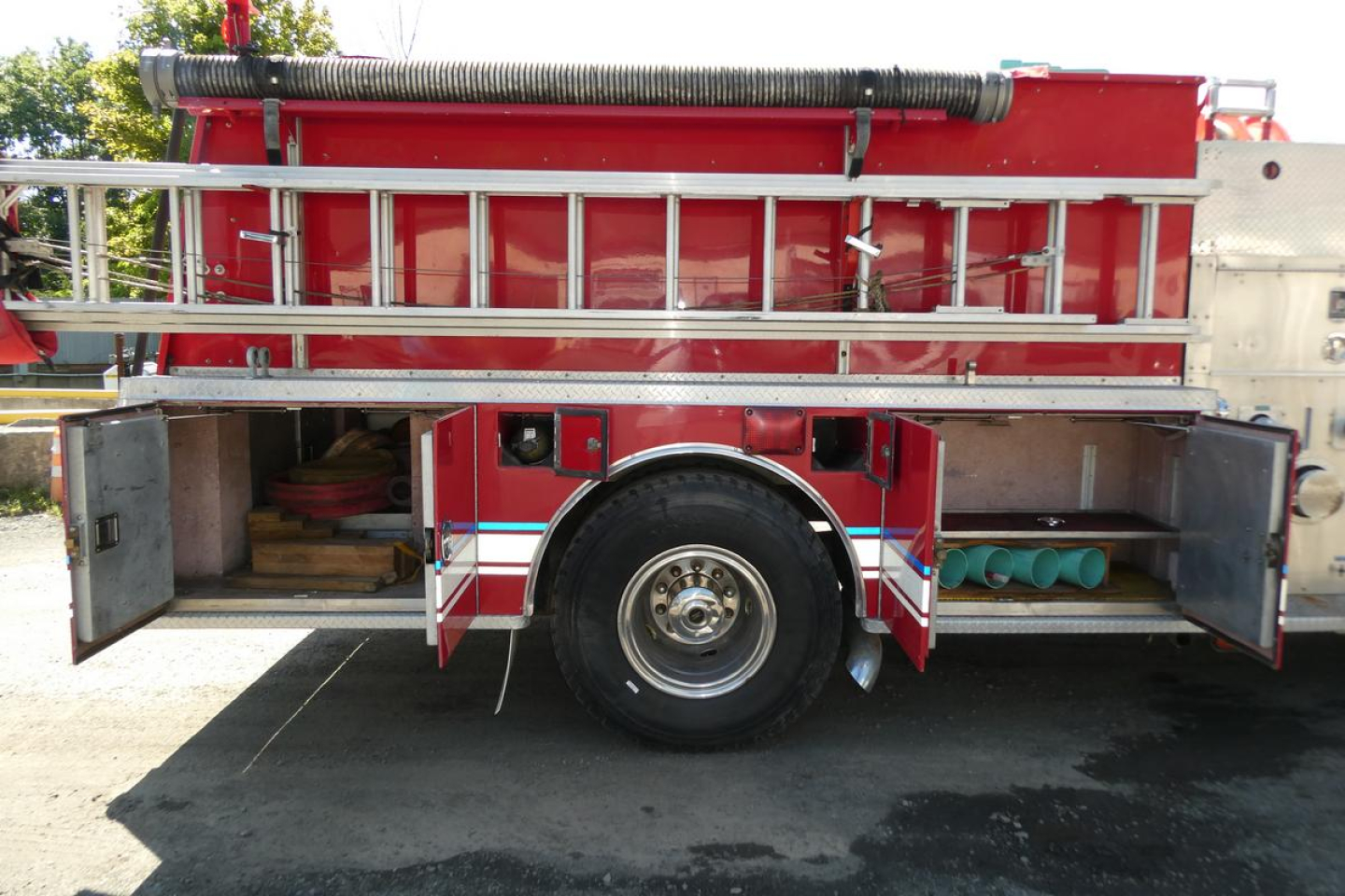 Fire truck side view showing open compartments and shelves with equipment, red and white body, ladder rack, and circular rear wheel.