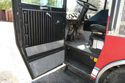 Fire truck cab interior with open door showing black ribbed door panel, metal diamond plate step, steering wheel, and dashboard controls.