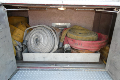 Truck compartment with coiled fire hoses and a nozzle, storage shelf, and metal floor; visible hoses include yellow and red colors.
