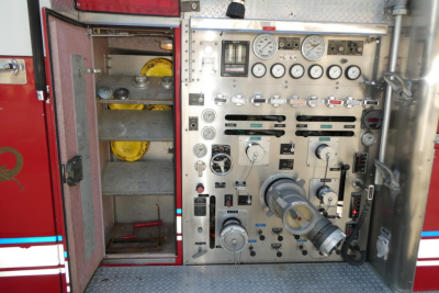 Fire truck pump panel with gauges, knobs, and hoses in an open compartment; shows control panel and yellow components inside a storage bay.