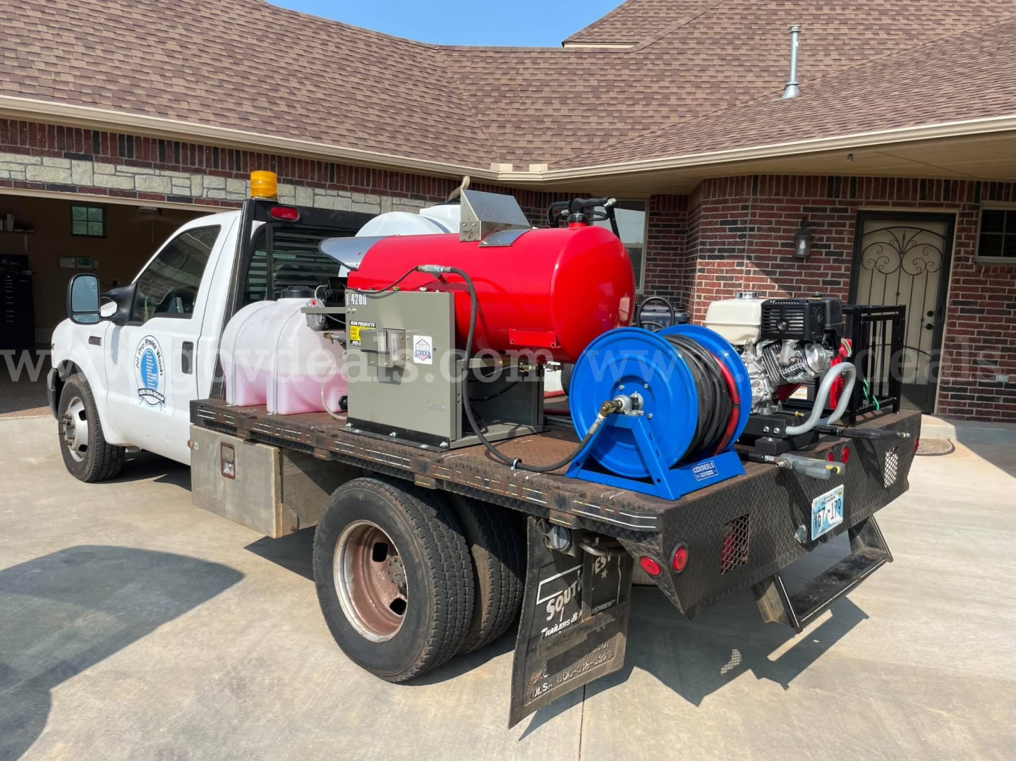 Flatbed truck with red tank, blue reel, and equipment mounted on the bed for service/utility use. Visible labels on equipment and hoses.