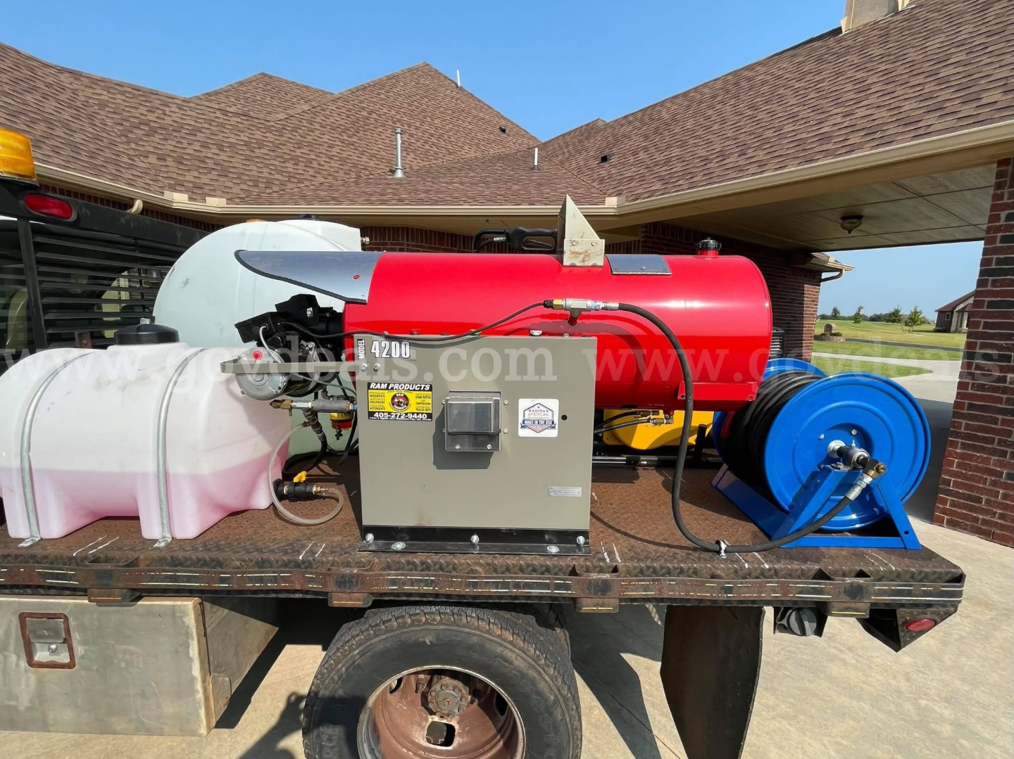 Truck bed with a red cylindrical pressure washer/tank and blue hose reel, gray generator box, and white/clear plastic tank mounted on a flatbed around a brick building.