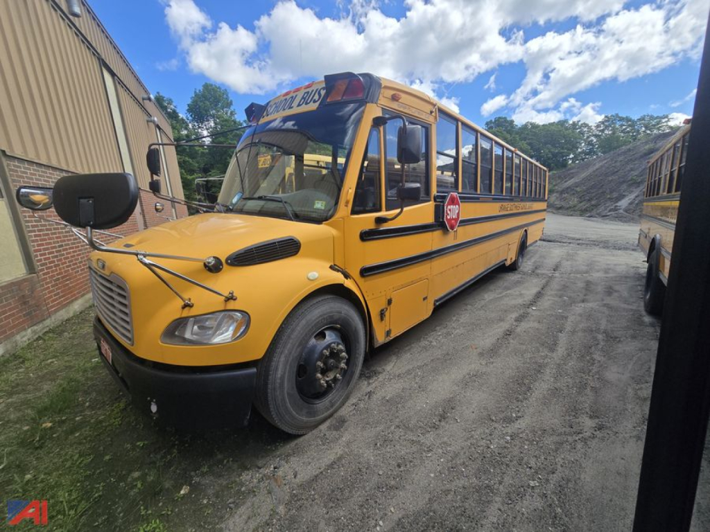 School bus in service yellow Freightliner bus with stop sign and bus logo on door, front view parked on gravel yard.
