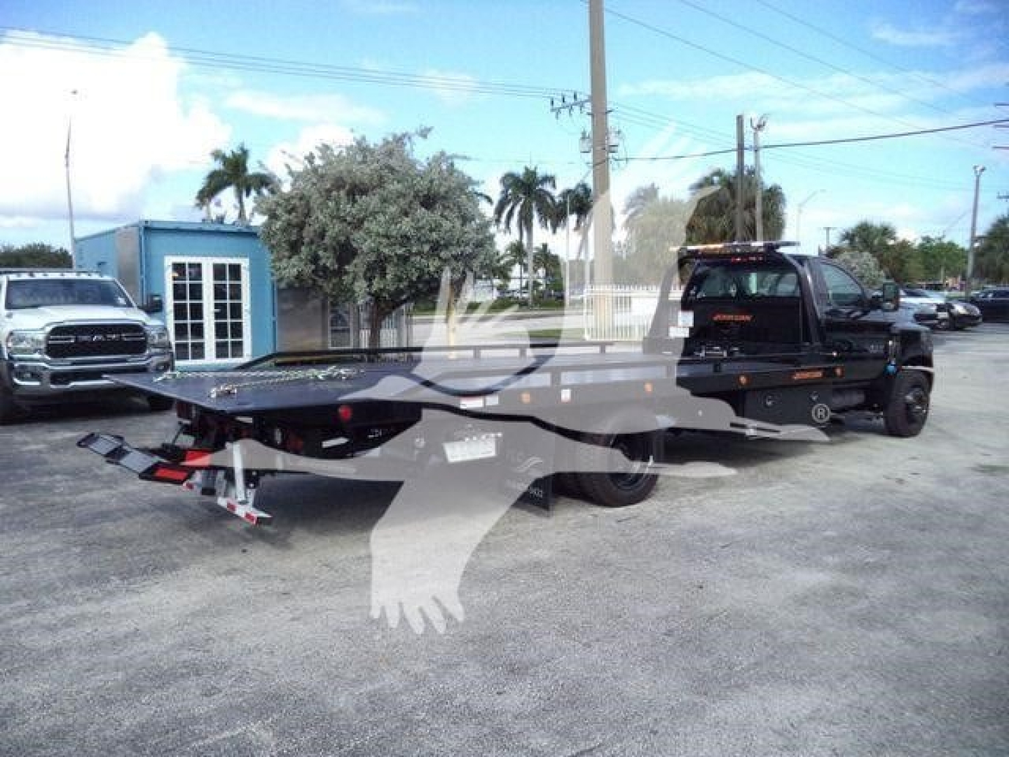 Flatbed rollback tow truck with a Chevrolet Silverado 6500HD chassis-cab in background, black pickup with equipment on bed, dealership lot setting.