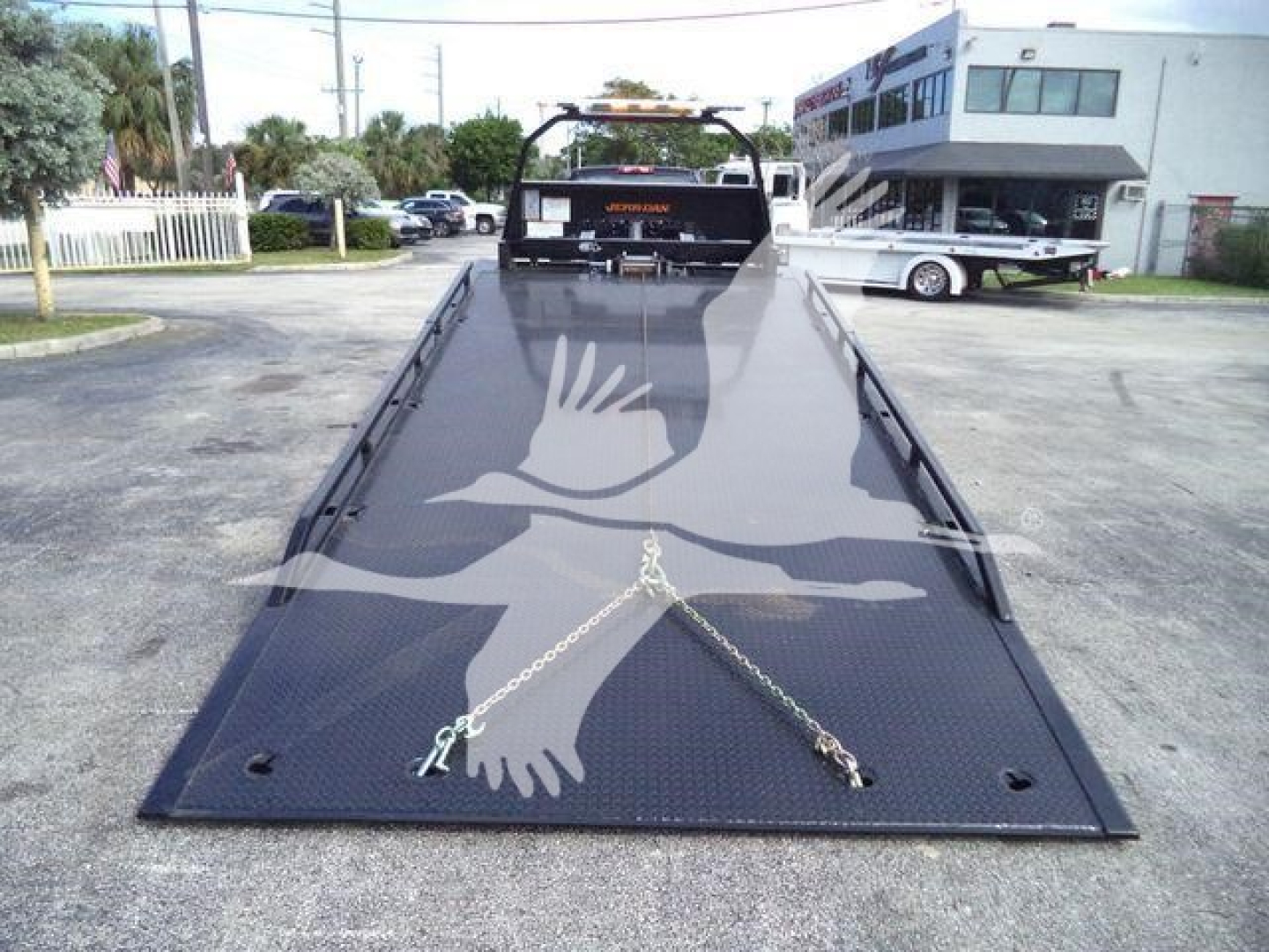 Truck bed of a 2023 Chevrolet Silverado 6500HD rollback with a black diamond-plate platform and ramp extending toward camera; chains securing front; dealership building in background.