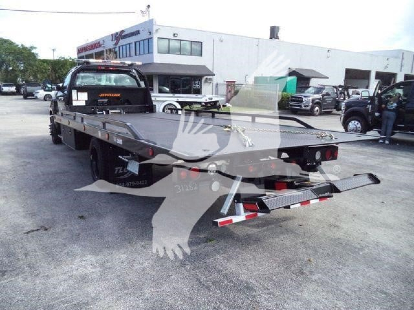 Flatbed tow truck with Silverado 6500 chassis shown from rear-right; aluminum bed, towing lights, ramps folded; dealership lot in background.