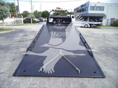 Truck bed of a 2023 Chevrolet Silverado 6500HD rollback with a black diamond-plate platform and ramp extending toward camera; chains securing front; dealership building in background.