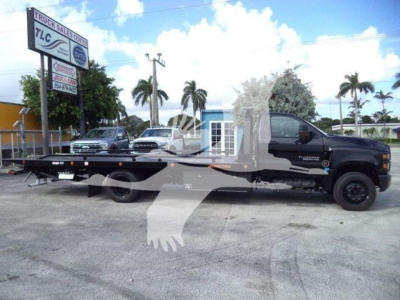 2023 Chevrolet Silverado 6500HD rollback truck in a dealership lot, black cab with long flatbed, dealership sign in background, stock number BLACK0UT20XLPFM.