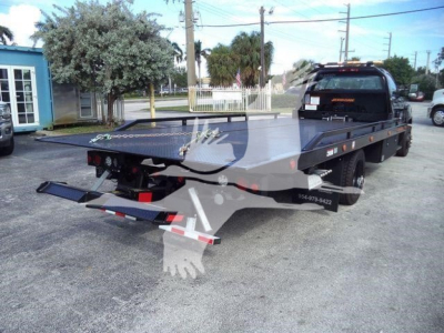 Flatbed tow truck with a Chevrolet Silverado 6500HD chassis in a dealership lot, rollback bed extended, black cab, rear view showing tow equipment and dual rear tires.