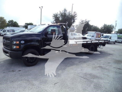 Truck: 2023 Chevrolet Silverado 6500HD Rollback with extended flatbed in a dealership lot; visible text includes Chevrolet emblem on grille and Silverado badge on door; multiple white vans in background.
