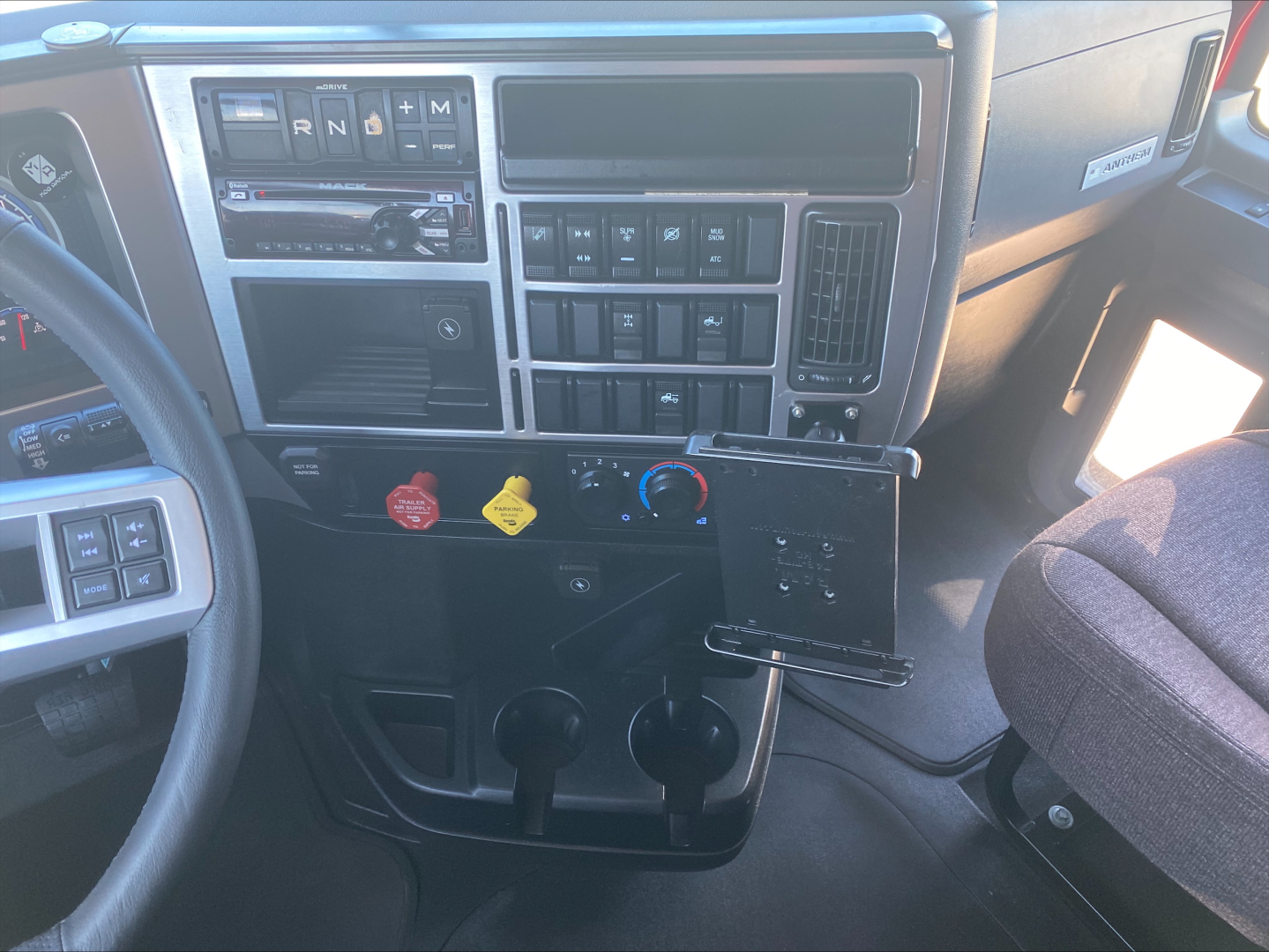 Dashboard of a 2020 Mack truck showing steering wheel, center console with gear indicators PRNDM, various switches, two cup holders, ignition area, red and yellow knobs, and a laptop or device mounted near the lower panel. Visible text incl