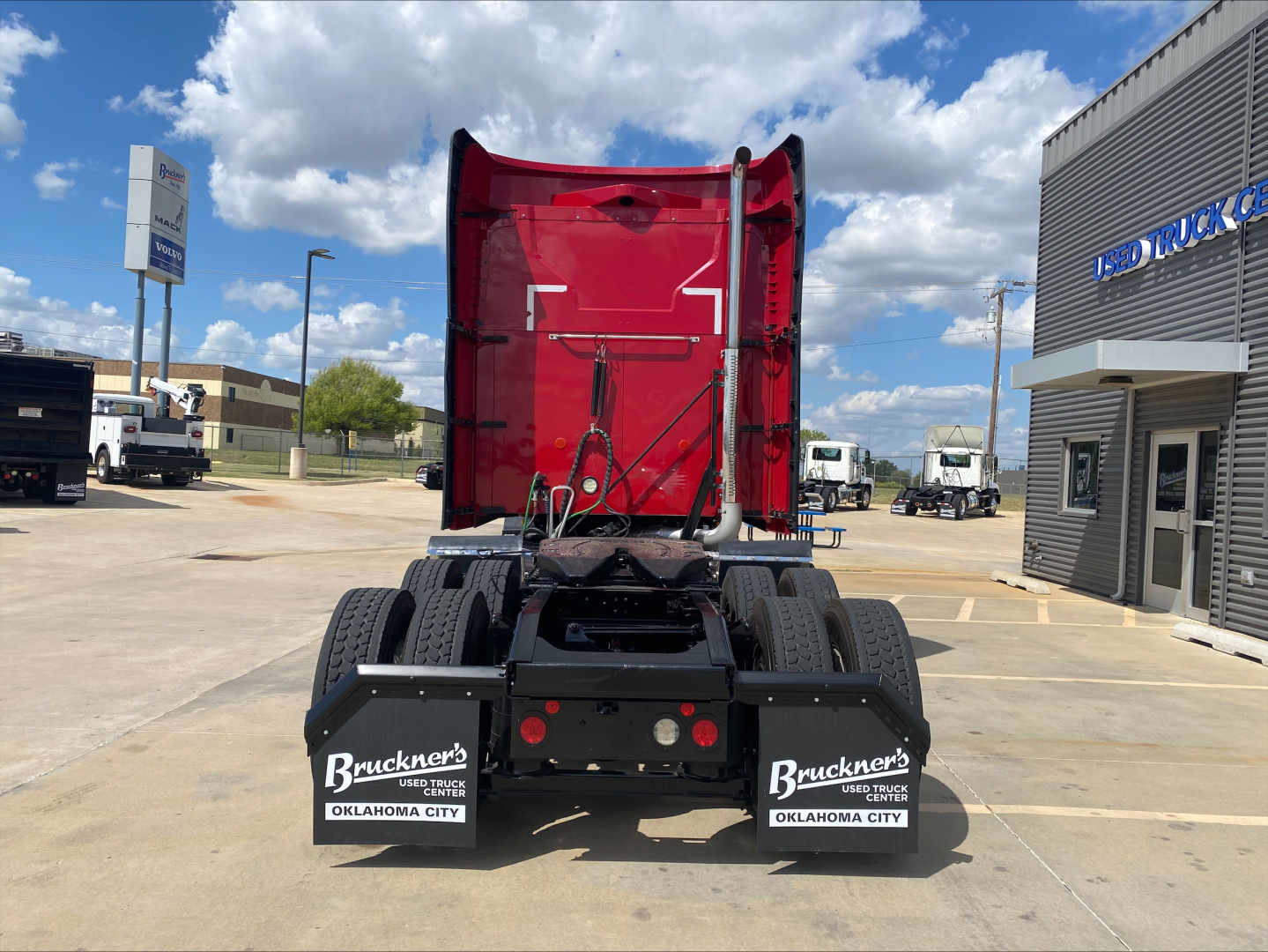 Rear view of a 2020 Mack AN64T70 tractor, red sleeper cab, black chassis, dual rear axles, Bruckner’s Oklahoma City mud flaps, dealership lot.