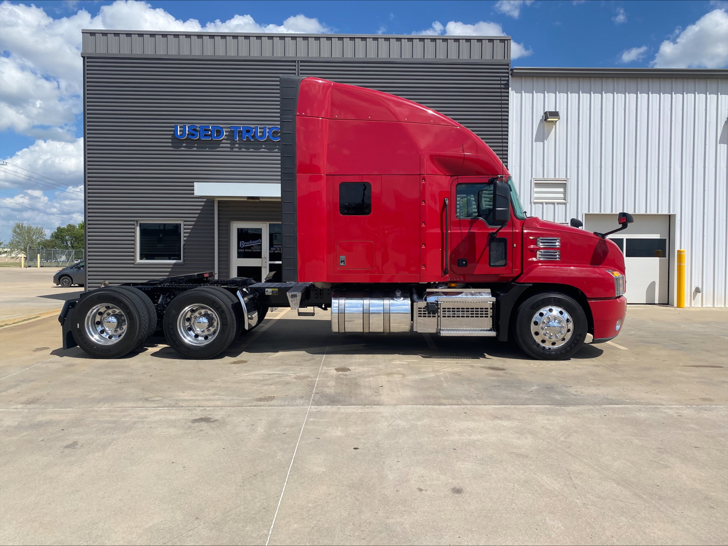 Red Mack AN64T70 truck tractor in front of a building with ‘USED TRUCK’ sign. Side view shows cab, sleeper with window, three rear axles, chrome fuel tanks and exhaust, and black tires on steel rims.
