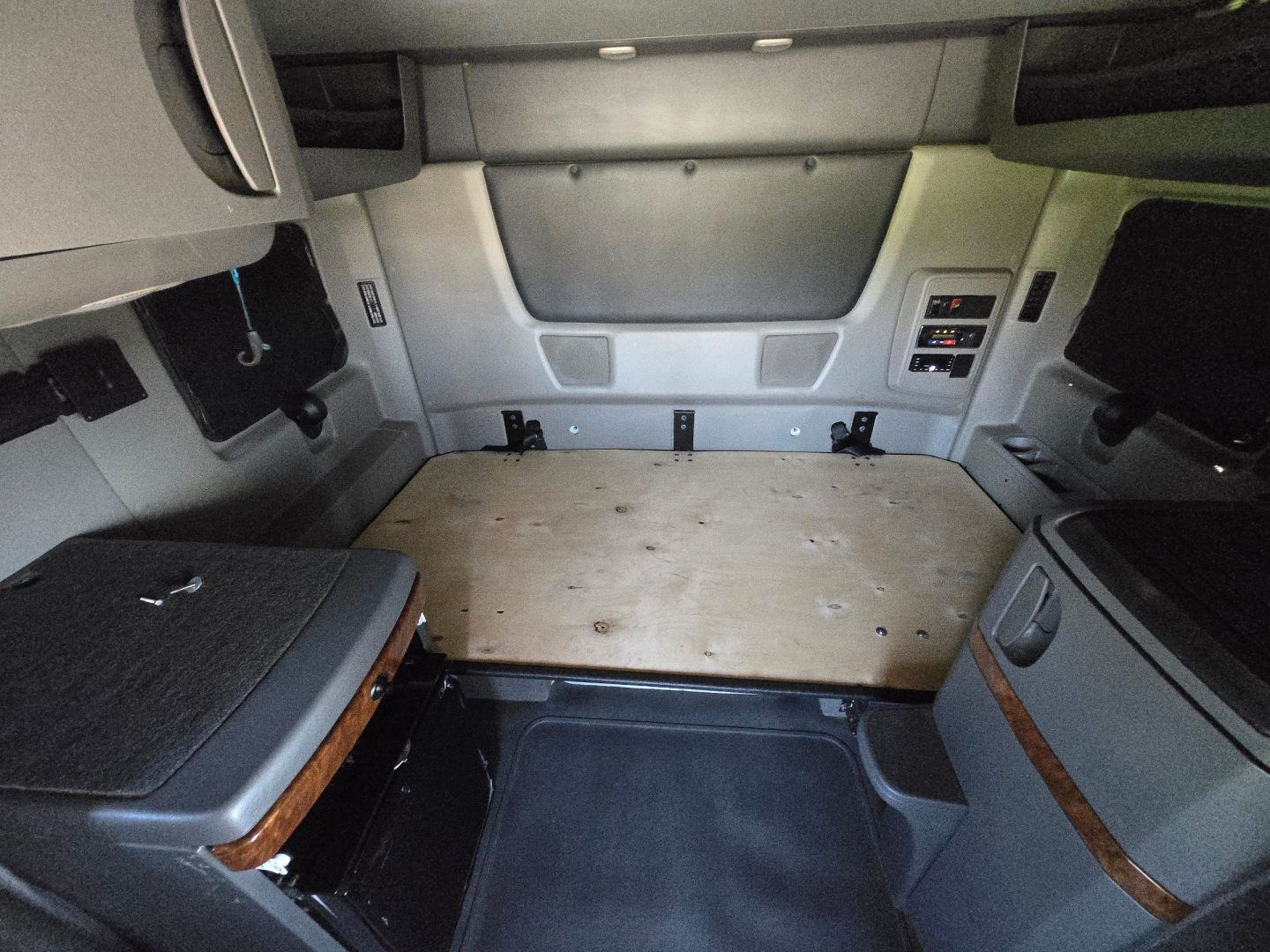 Truck cab interior showing sleeper area with plywood floor and side panels; dashboard and control modules visible on right side.