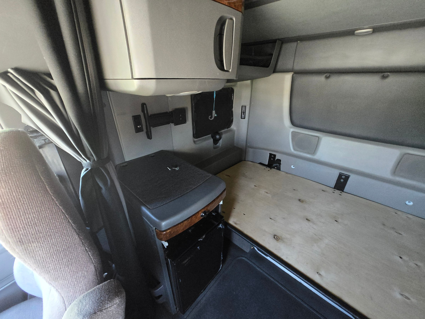Cab interior of a 2021 International LT625 sleeper truck showing the driver seat, overhead storage, and a plywood platform over the sleeper area.