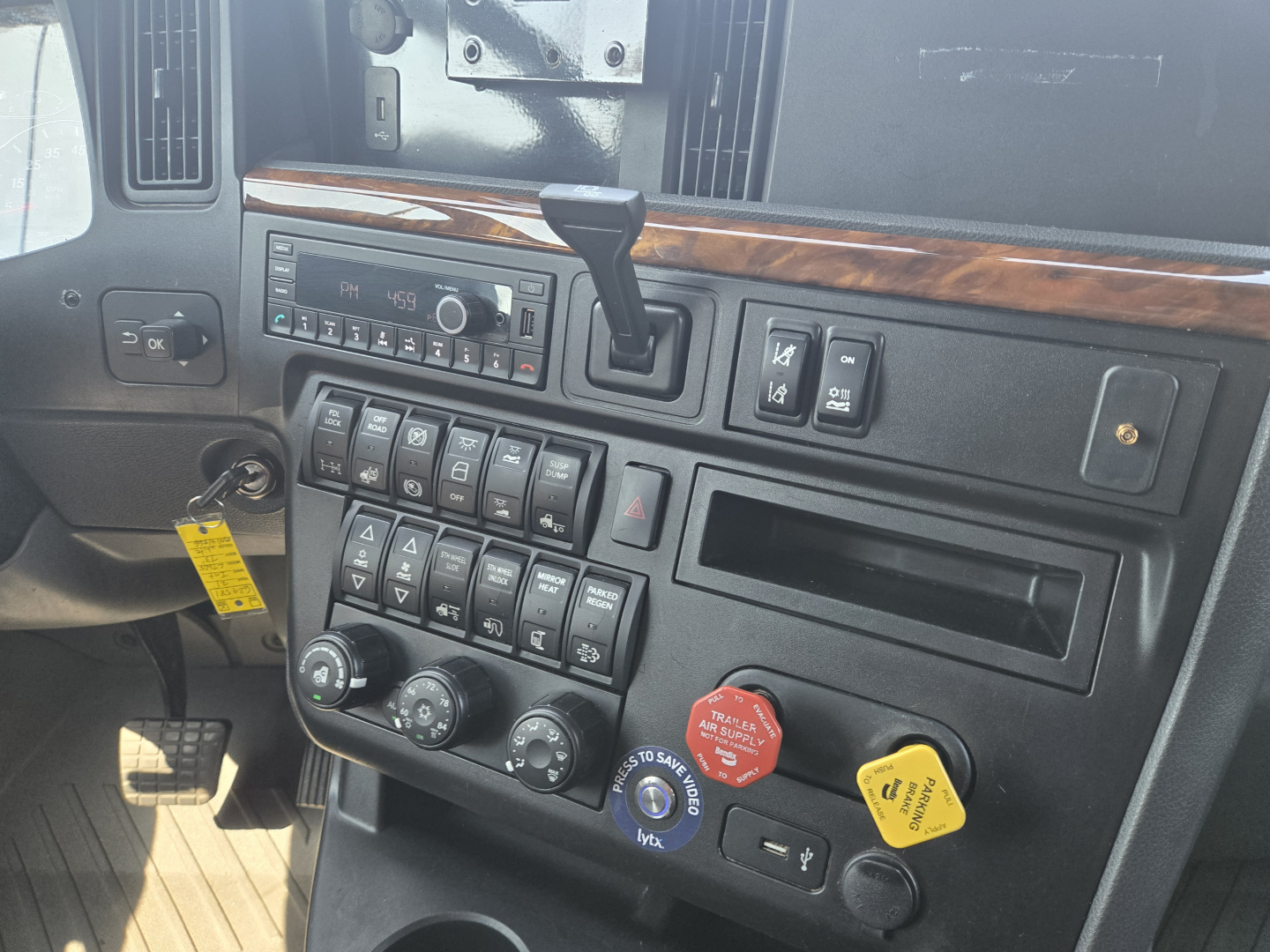 Dashboard area of a 2021 International LT625 truck showing steering column, climate controls, many toggle switches, and a red safety cap; some wood dash trim and a radio display visible.