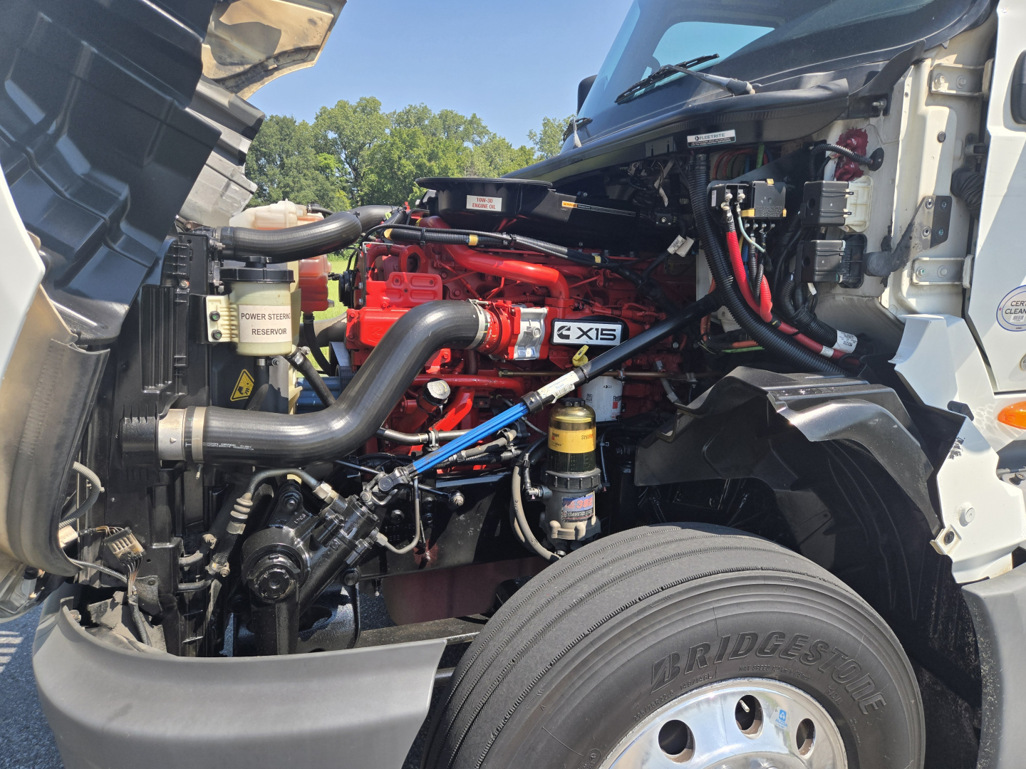 Engine bay of a 2021 International LT625 truck showing red engine block and intake, various hoses, tubing, and a Bridgestone front tire in foreground.