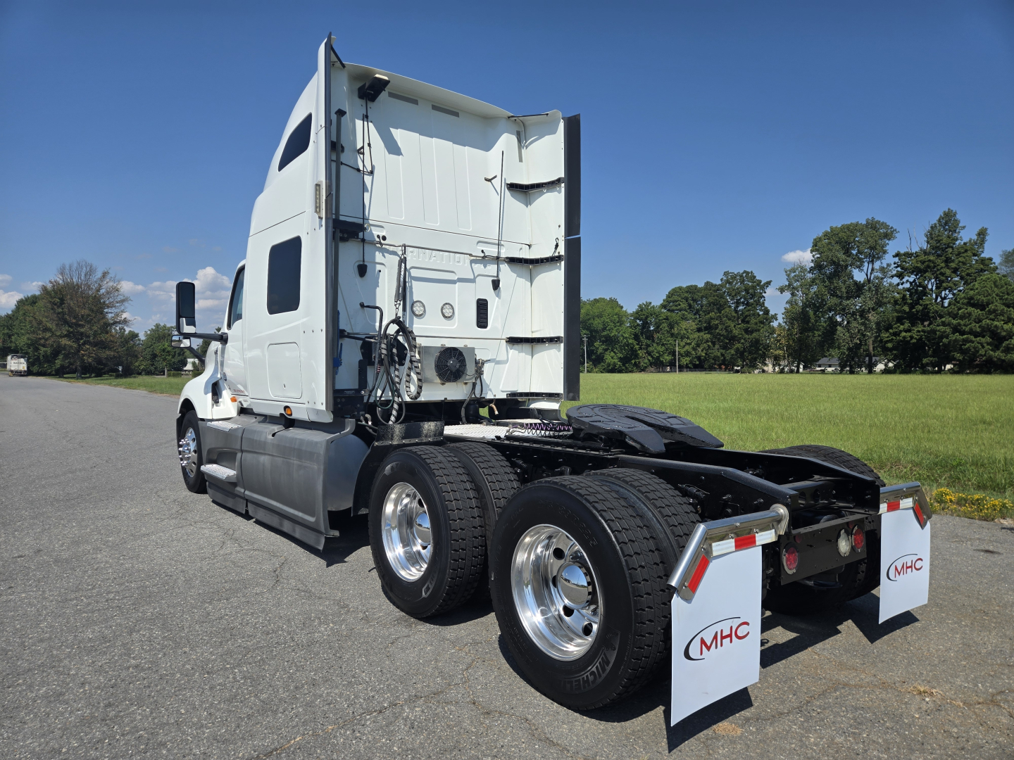 Truck rear-left view showing dual rear axles, fifth-wheel area, white sleeper cab, and mud flaps with MHC logo.
