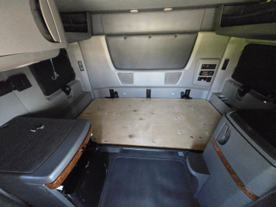 Truck cab interior showing sleeper area with plywood floor and side panels; dashboard and control modules visible on right side.