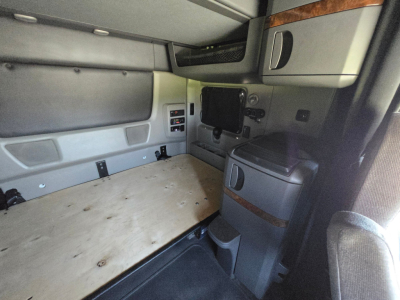 Truck cab interior showing sleeper area with wooden floor panel, side cabinets, control panel, and a mounted screen; grey and black trim with storage compartments.