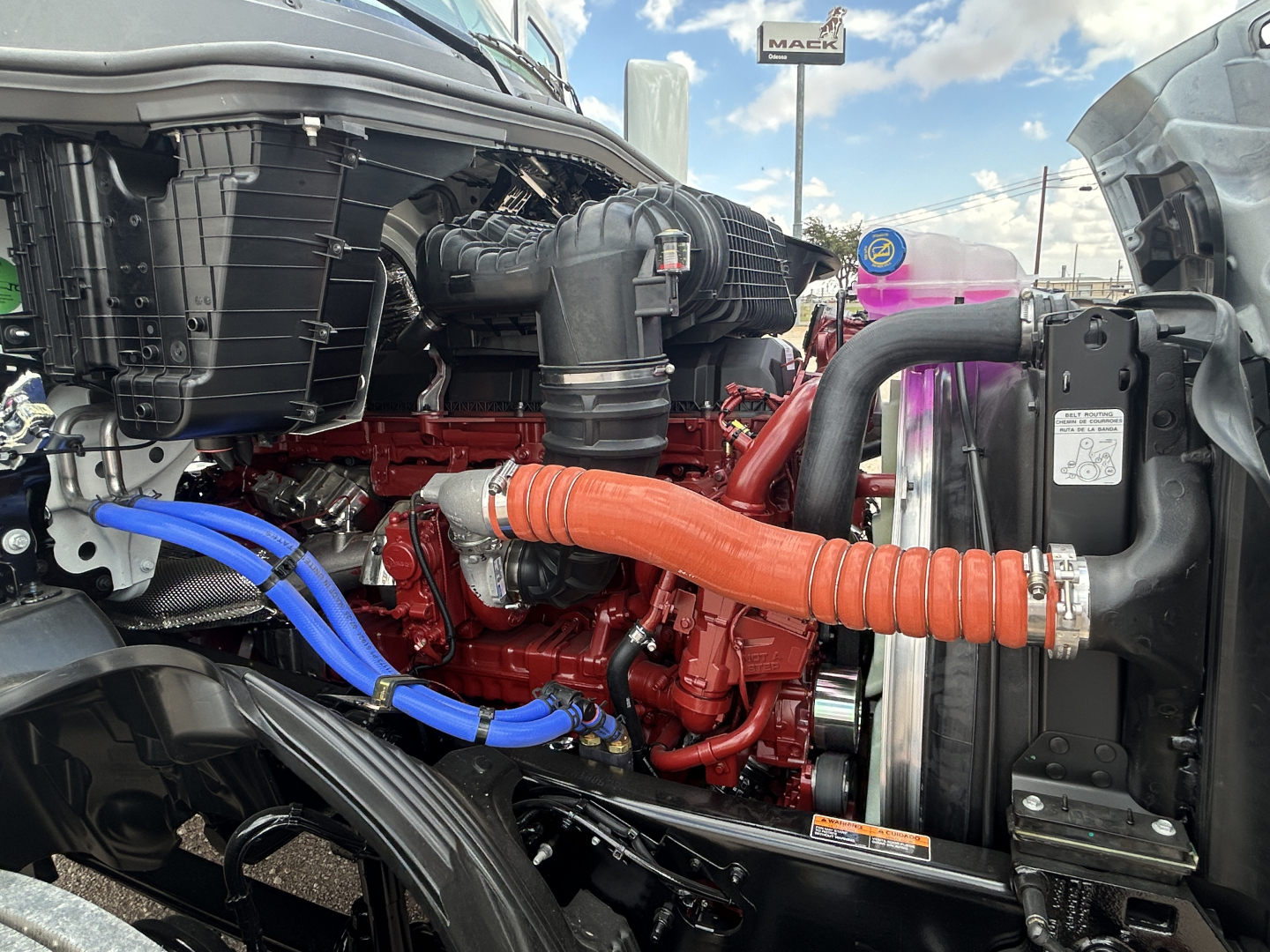 Mack PR64T engine bay showing red engine block with orange flexible hose connecting to radiator and blue hoses, front grille area visible.