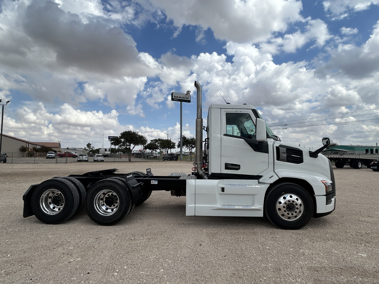 Front half view of a white 2026 Mack PR64T conventional daycab truck, side profile exposed with triple rear axle, no trailer, clear sky.