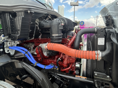 Mack PR64T engine bay showing red engine block with orange flexible hose connecting to radiator and blue hoses, front grille area visible.