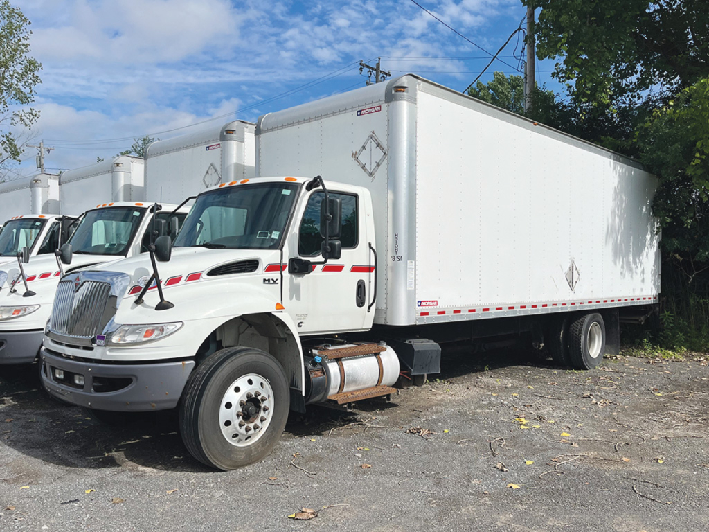 White International MV607 conventional daycab truck with a large white trailer, front left view, stock numbers and VIN notations visible.