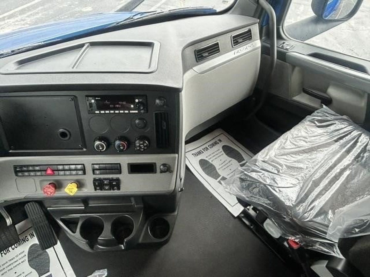 Truck dashboard interior of a 2022 Freightliner Cascadia 126 with grey cabin, steering area, center console, and plastic-covered seat.