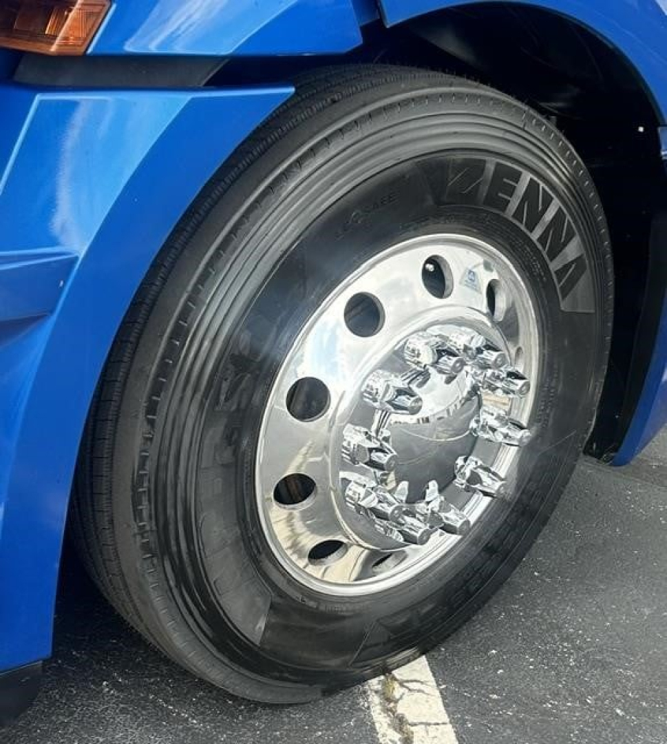 Front left wheel of a blue Freightliner Cascadia 126 with Zenna tire brand and chrome hubcap; partial fender and headlight visible.