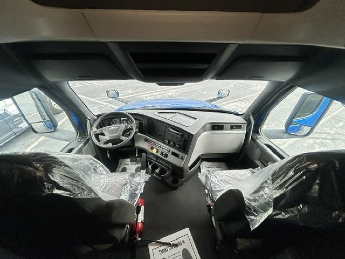Truck cab interior of a 2022 Freightliner Cascadia 126 from the driver’s perspective, with plastic covers on seats, large dashboard, steering wheel, and center console.