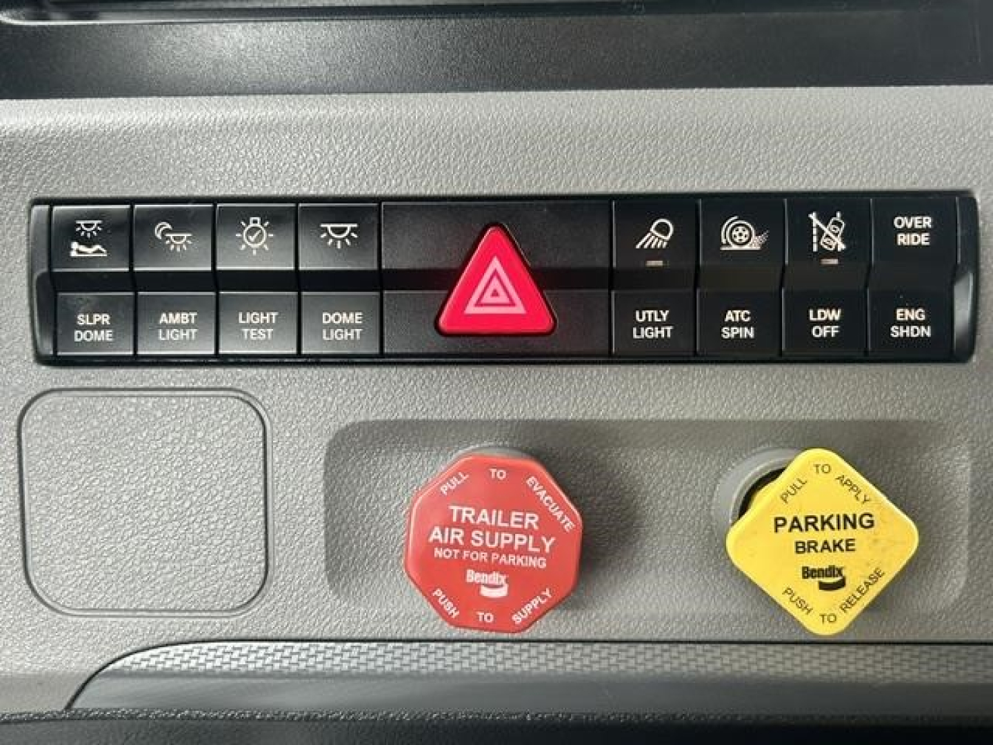 Dashboard switch panel and two colored caps (red and yellow) in a Freightliner Cascadia cab. Red cap labeled TRAILER AIR SUPPLY and yellow cap labeled PARKING BRAKE. Various buttons above include hazard light and utility lights.
