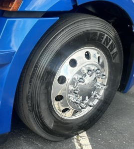 Front left wheel of a blue Freightliner Cascadia 126 with Zenna tire brand and chrome hubcap; partial fender and headlight visible.