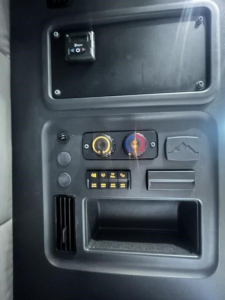 Cab interior panel of a Freightliner Cascadia showing dashboard controls and circular dials with yellow and red rings, a rectangular storage slot, and ventilation grille.