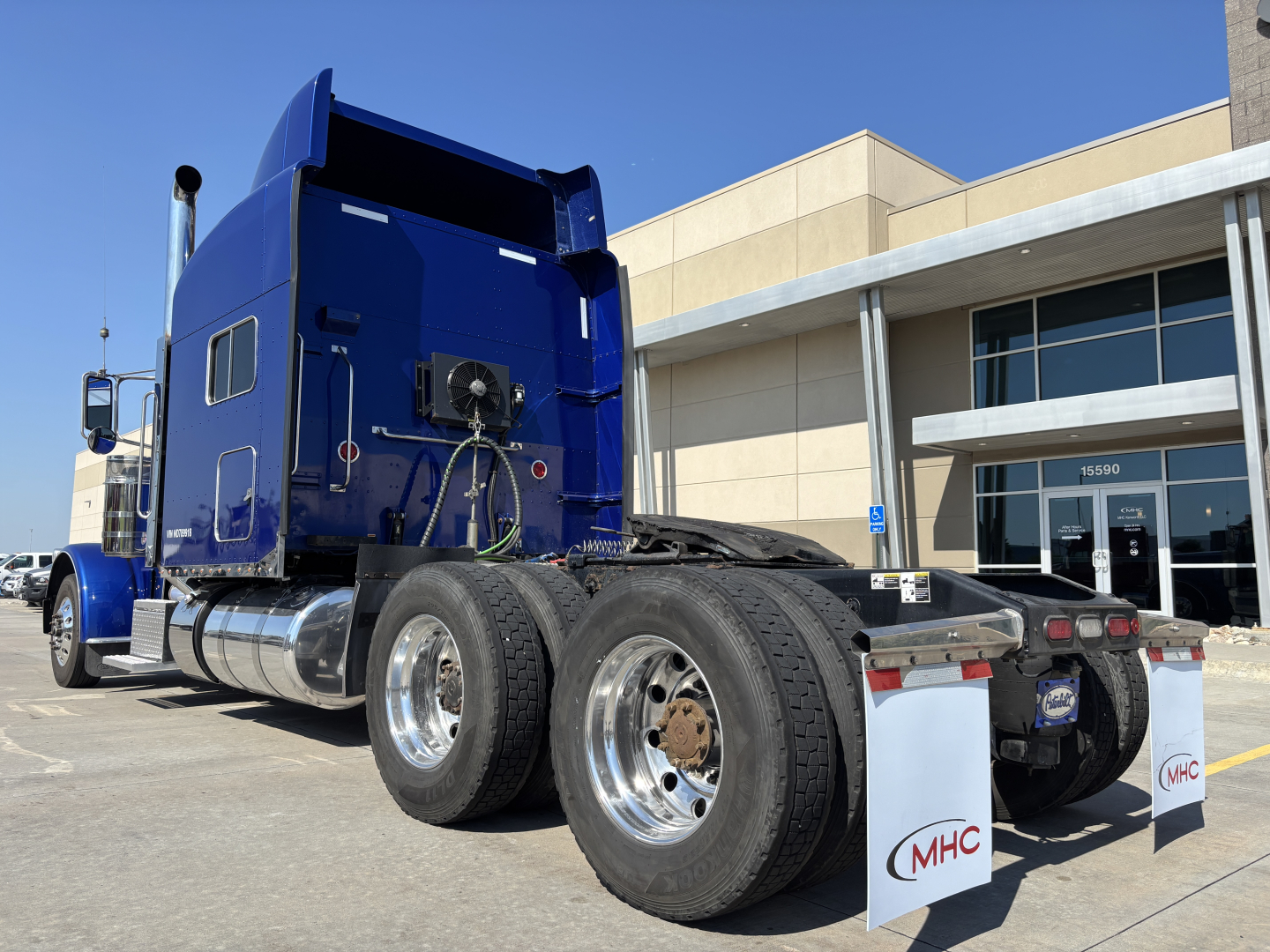 Peterbilt 389 dual rear wheels tractor cab in blue, side view, with sleeper and chrome fuel tanks. VIN tag not visible, blue dealership building in background.