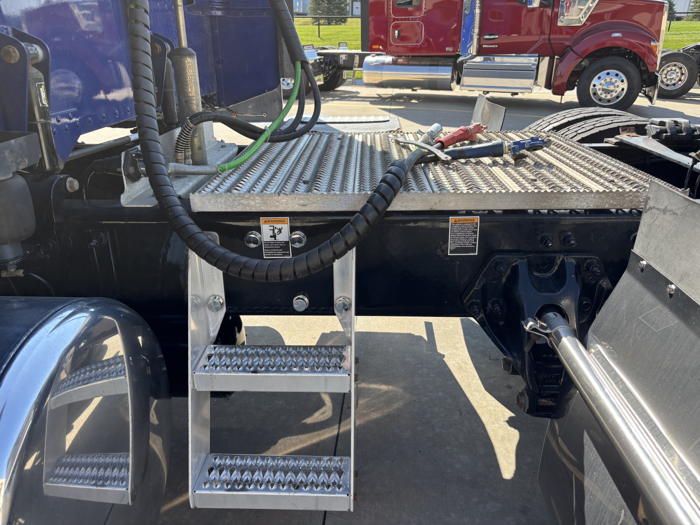 Peterbilt 389 engine bay area showing metal deck with steps, ladder, and hydraulic lines; metal fuel/air lines and a chrome hitch shaft visible under cab, utility cables across the platform