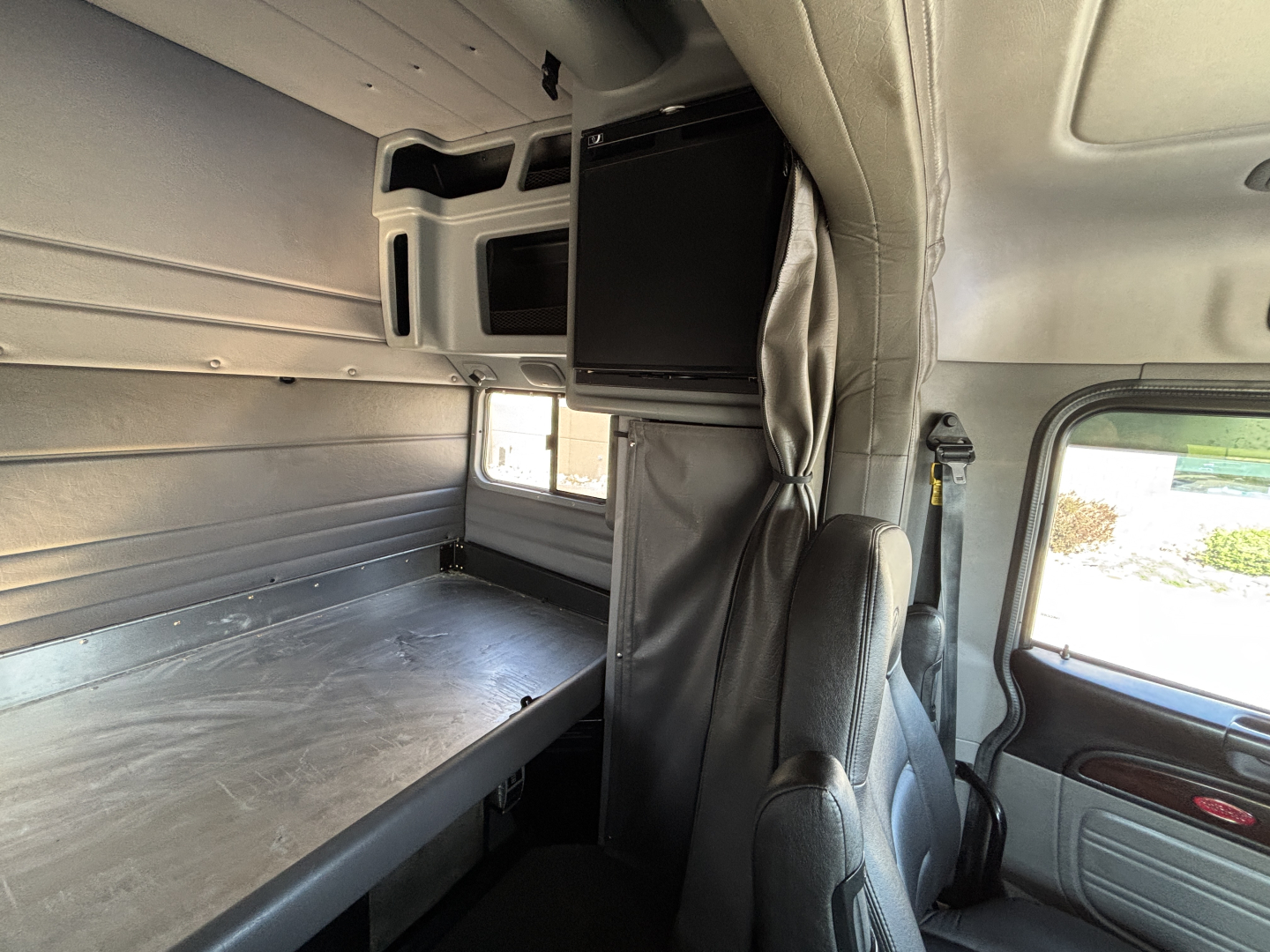 Peterbilt truck interior sleeper cab with a vertical TV mounted and bunk area along left wall; captain seat in foreground and divider curtain.