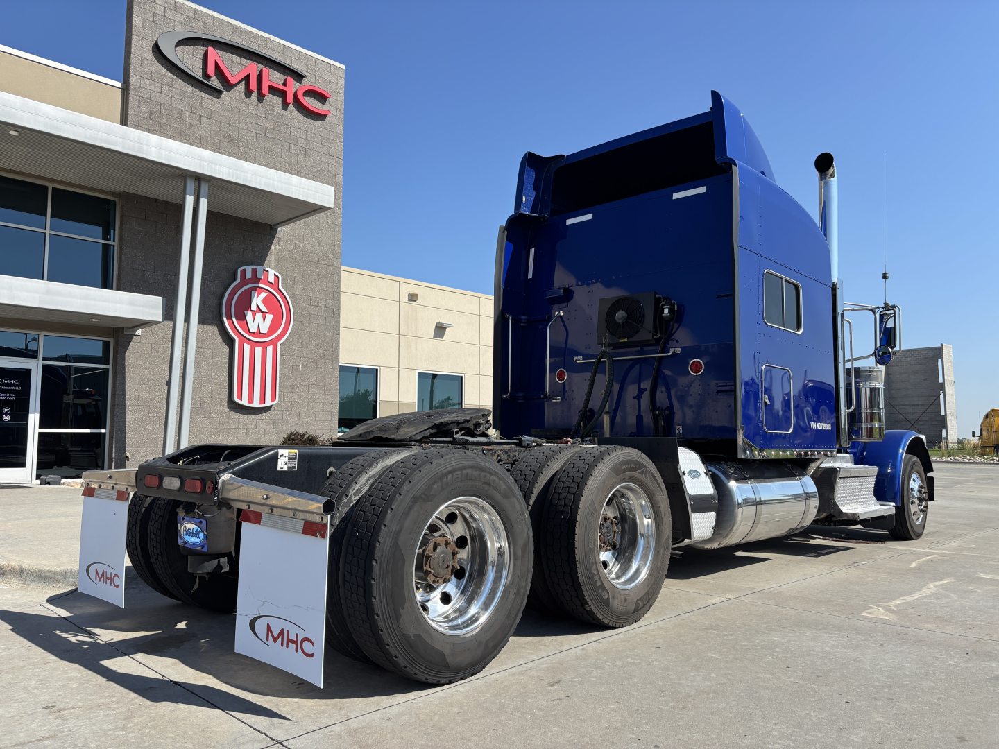 Peterbilt 389 tractor in blue with sleeper cab, dual rear axles, photographed from rear side showing chrome fuel tanks and mud flaps.