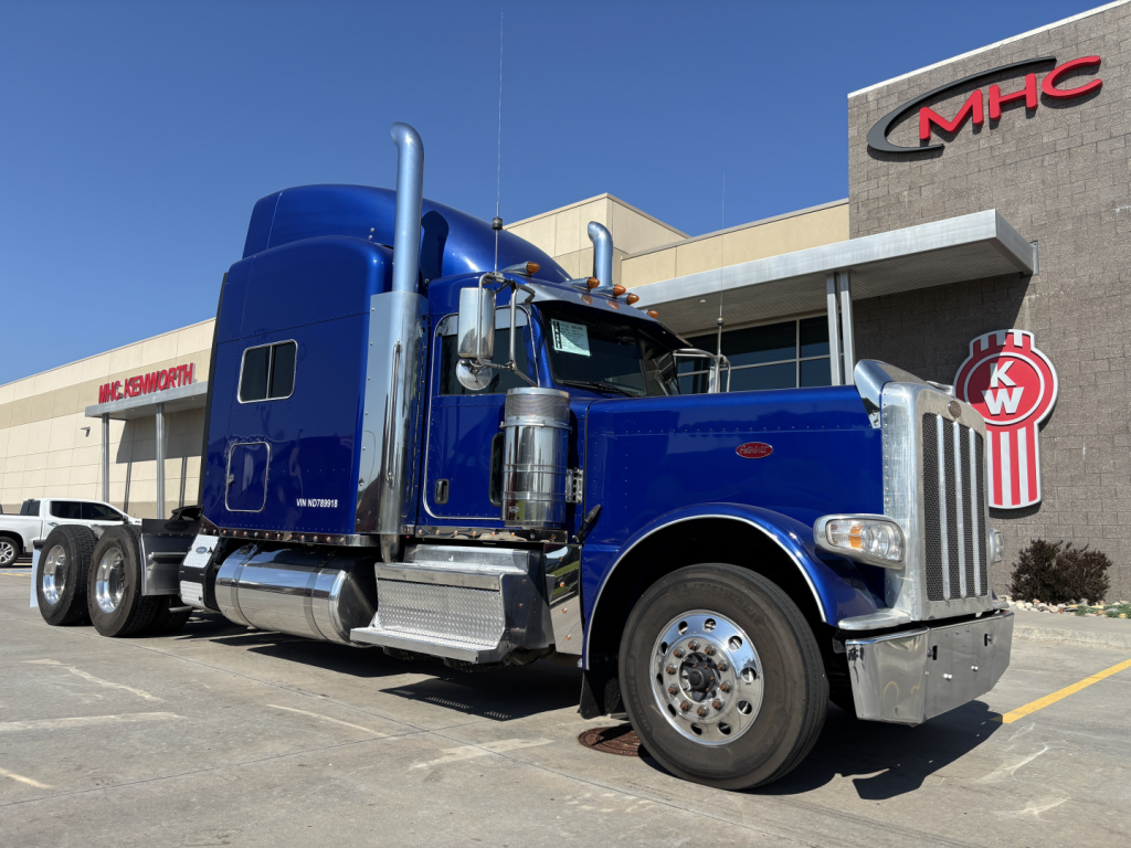 Peterbilt 389 tractor in blue, side view showing chrome exhaust stacks, sleeper cab, dual rear axles, and chrome fuel tanks in front of dealership building.