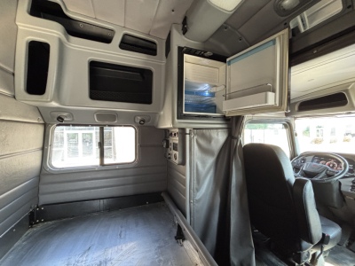 Peterbilt 389 sleeper cab interior showing open overhead cabinet with refrigerator, driving seat, and cargo area. Cabin walls with light panels and storage compartments.