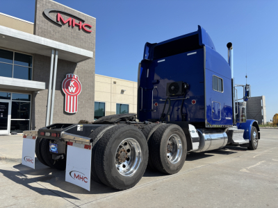 Peterbilt 389 tractor in blue with sleeper cab, dual rear axles, photographed from rear side showing chrome fuel tanks and mud flaps.
