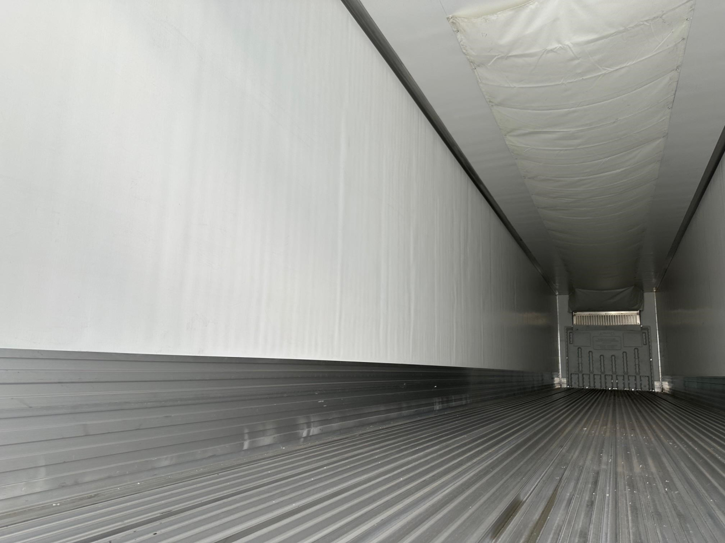 Trailer interior showing long empty refrigerated trailer with ribbed floor and white insulated walls, rear doors visible at far end.