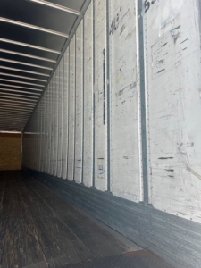 Interior of a cargo trailer showing aluminum side walls with vertical seams and a dark wooden floor.