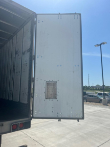 Rear door of a large cargo trailer open, showing the interior wall with a small vent/vent-like panel and riveted metal panels on the door.