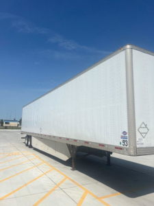 Trailer side view showing a white utility trailer with two axles, front corner post, and small decals near the front corner. Clear blue sky.