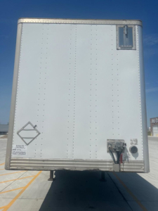 Rear view of a white utility van trailer showing a diamond placard, rear bumper riser, and electrical connection box with cables.