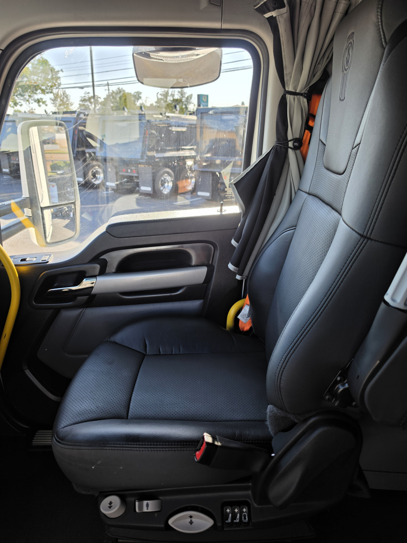 Truck cab seat interior showing driver seat and door panel of a Kenworth T680 conventional sleeper, with window view of other trucks outside.