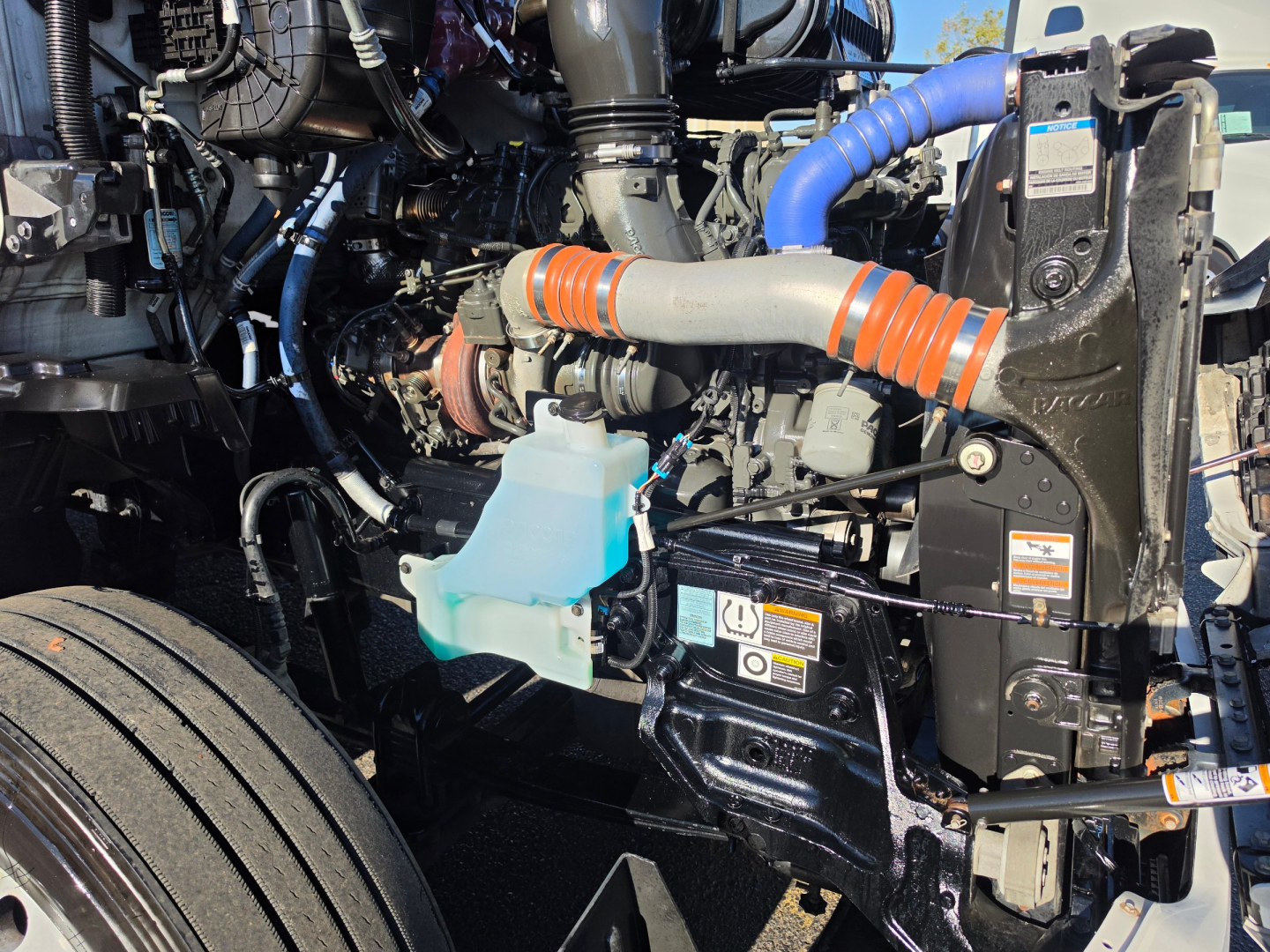 Kenworth T680 engine bay showing coolant reservoir, orange helical hose, blue radiator hose, and various engine components.