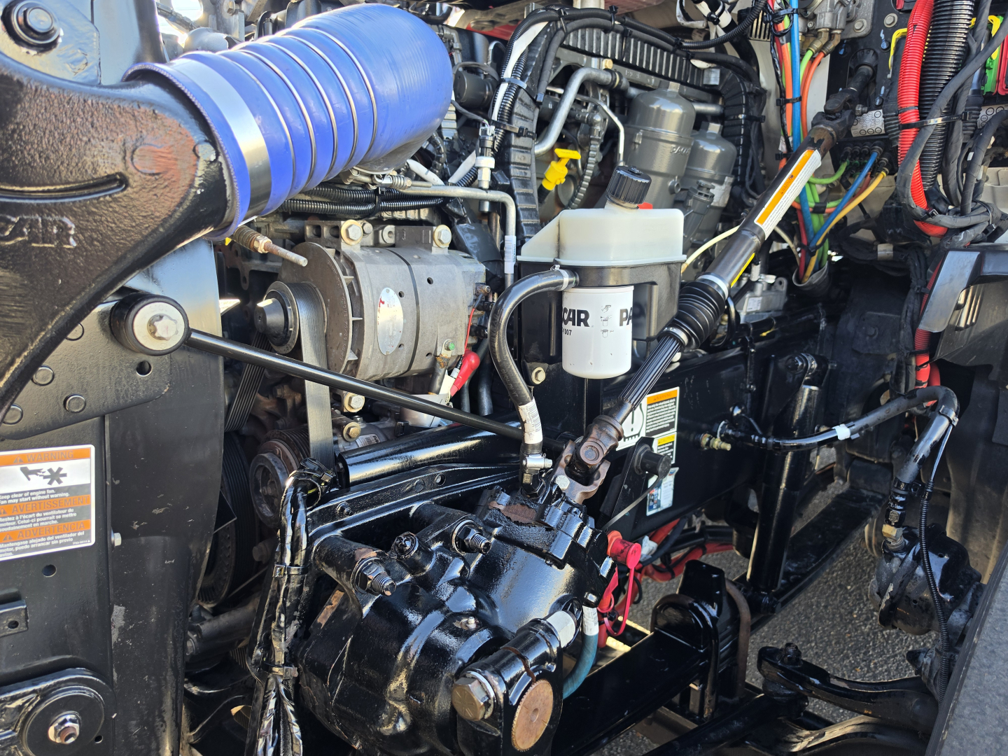 Kenworth T680 engine bay view showing engine components, hoses, and reservoir; visible white brake reservoir with black cap and lines; various cables and components including a blue intake hose.