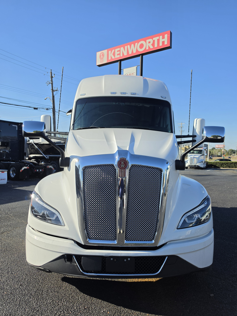 Kenworth T680 front view showing large chrome grille, headlights, and Kenworth dealership sign in background.